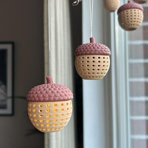 Könnte beinhalten: Ein Set aus hängenden eichelförmigen Ornamenten, die an einem Ast befestigt sind. Jedes Ornament hat eine texturierte, malvenfarbene Kappe und eine perforierte, hellbraune Basis, die von innen warmes gelbes Licht ausstrahlt und sanft leuchtet.