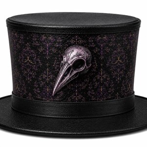 Op de afbeelding: Een zwarte hoge hoed met een gestructureerde, leerachtige rand en kroon, versierd met een paars en zwart damastpatroon aan de zijkant. Een metalen paars vogelschedel-embleem is in het midden aan de voorzijde van de hoed bevestigd.