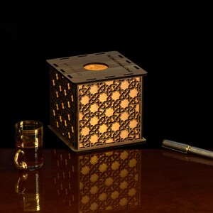 May include: A square wooden lamp with intricate laser-cut geometric lattice patterns on its sides, emitting a warm internal glow. The top features a circular opening. A small glass with a gold rim and a silver pen are placed nearby on the surface.