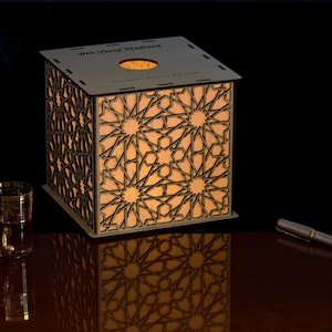 May include: A square wooden lamp with intricate laser-cut geometric patterns on its sides, emitting a warm internal glow. The top surface features a circular cutout and the text Ben Youssef Madrasa. A glass and a pen are placed nearby.