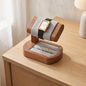May include: A wooden watch and ring display stand featuring a curved, light gray velvet-lined holder for a gold-cased wristwatch with a black textured strap, and a base with two velvet-lined slots containing two plain gold-toned metal rings.