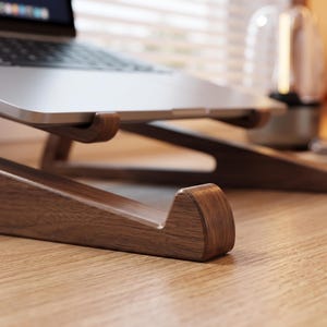 Puede incluir: Un soporte para portátil de madera marrón oscuro con un acabado liso y pulido y un borde de apoyo frontal curvado. El soporte presenta un diseño en ángulo para elevar un portátil plateado, mostrando la textura natural de la veta de la madera.