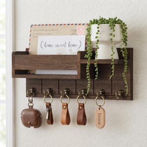 May include: A dark brown wooden wall-mounted organizer featuring a mail slot, a small shelf with a white planter containing a trailing green plant, and six brass hooks. Several keychains and a brown case hang from the hooks, and an envelope is in the slot.