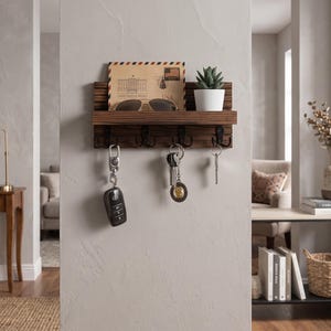 May include: A dark brown wooden wall-mounted shelf featuring four black metal hooks for hanging items. The shelf holds a tan envelope with airmail markings, a pair of sunglasses, and a small white pot containing a green succulent plant.