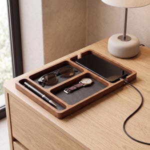 Puede incluir: Una bandeja organizadora rectangular de madera con cuatro compartimentos distintos y un forro interior gris oscuro. La bandeja contiene un teléfono inteligente negro conectado a un cable, gafas de sol, un reloj con correa marrón y un bolígrafo.