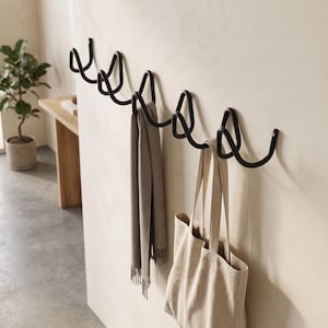 Op de afbeelding: Een matzwarte wandkapstok bestaande uit een doorlopende, golvende metalen draad met meerdere haken. Een grijze sjaal met ruitpatroon en een beige canvas tas hangen aan de haken van dit functionele wandopbergsysteem voor aan de muur.