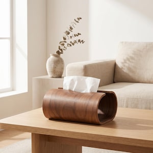 Puede incluir: Una funda para caja de pañuelos cilíndrica y horizontal hecha de madera de color marrón oscuro con un patrón de veta visible. La funda presenta una abertura rectangular larga y estrecha en la parte superior, por donde se extrae un pañuelo blanco.