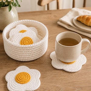 May include: A set of white and yellow crocheted coasters shaped like fried eggs, with several stored in a round white crocheted basket and one placed under a speckled ceramic mug. The coasters feature a central yellow circle and white edges.