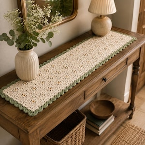 May include: A rectangular crochet table runner featuring a repeating cream-colored floral motif pattern with a scalloped sage green border. The textile is crafted with intricate yarn work, showcasing a delicate openwork design throughout the piece.