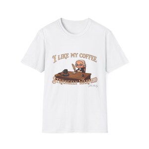 May include: A white short-sleeve t-shirt featuring a graphic of a cartoon figure sitting at a wooden desk with a quill and coffee mug. The text reads I LIKE MY COFFEE STRANGELY WARMED and includes the signature John Wesley below the desk.