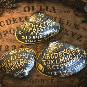 Puede incluir: Tres conchas marinas negras pintadas con letras doradas que recuerdan a tableros Ouija. Cada concha muestra el alfabeto, los números del cero al nueve y las palabras YES, NO, HELLO y GOODBYE. Acabado brillante y bordes amarillos.