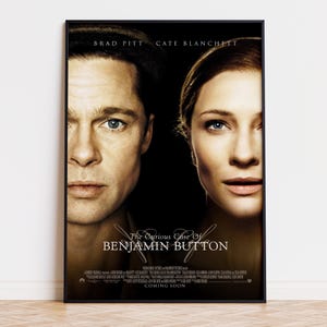 May include: A framed movie poster for The Curious Case of Benjamin Button featuring close-up portraits of Brad Pitt and Cate Blanchett against a dark background. The title is printed in white text, with additional production credits at the bottom.