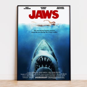Pode incluir: Um cartaz de filme Jaws emoldurado apresentando um grande tubarão nadando em direção à superfície onde uma pessoa nada. O título JAWS está em letras vermelhas em negrito no topo. O texto inclui nomes de atores, créditos de produção e o slogan.
