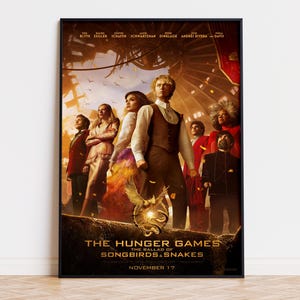 Op de afbeelding: Een filmposter voor The Hunger Games: The Ballad of Songbirds & Snakes met een groep personages in formele kleding tegen een warme, goudkleurige achtergrond met een centraal embleem van een vogel en slang, en de datum 17 november.
