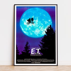 Puede incluir: Un póster de película enmarcado de E.T. El Extraterrestre que muestra la silueta de una persona en bicicleta volando frente a una gran luna llena azul brillante. El fondo es un cielo nocturno de color púrpura oscuro con siluetas de árboles.