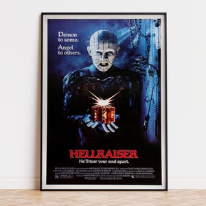 May include: A framed Hellraiser film poster featuring the character Pinhead with pins in their head, holding a glowing puzzle box. The text reads: Demon to some. Angel to others. HELLRAISER He'll tear your soul apart. The background is dark blue.