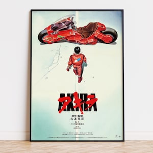 Puede incluir: Un póster enmarcado que muestra una motocicleta roja y una figura con chaqueta roja caminando. El texto AKIRA aparece en letras negras grandes con detalles rojos, seguido de caracteres japoneses y créditos sobre un fondo claro.