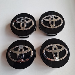 Puede incluir: Un juego de cuatro tapacubos circulares, cada uno con un acabado negro brillante y un emblema de Toyota cromado y elevado en el centro. Estos accesorios para automóviles están dispuestos en una cuadrícula de dos por dos.
