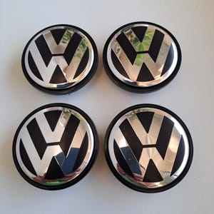 Peut inclure: Un ensemble de quatre enjoliveurs de roue circulaires arborant le logo Volkswagen avec une finition chrome poli sur un fond noir mat. Chaque enjoliveur présente un contour noir et une surface métallique réfléchissante formant l'emblème VW.