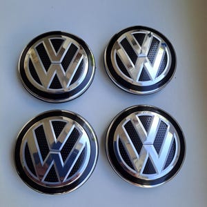Peut inclure: Un ensemble de quatre enjoliveurs de roue circulaires Volkswagen arborant le logo VW en finition chromée sur un fond noir perforé. Chaque enjoliveur présente une bordure argentée polie et un design moderne aux lignes métalliques nettes.