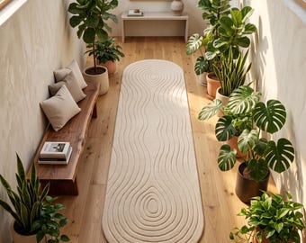May include: An elongated oval-shaped area rug featuring a cream-coloured surface with a textured, concentric ripple pattern. The rug is positioned on a light-toned wooden floor in a hallway flanked by potted green plants and a wooden bench.
