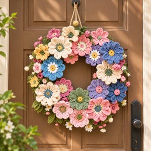 Puede incluir: Una corona circular hecha de flores de ganchillo en tonos rosa, azul, amarillo, crema y verde. El arreglo floral presenta varias formas y tamaños de pétalos, con pequeñas hojas de ganchillo verdes intercaladas en el diseño.