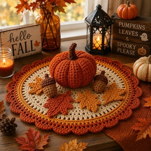 May include: A round, crocheted table mat in cream and orange with a scalloped edge, featuring a large orange crocheted pumpkin, two small brown crocheted acorns, and three crocheted autumn leaves in shades of orange and brown.