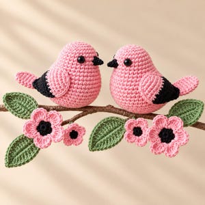 Könnte beinhalten: Zwei gehäkelte Vögel mit rosa Körpern, schwarzen Flügeln und schwarzen Schnäbeln sitzen auf einem braunen Ast. Der Ast ist mit gehäkelten grünen Blättern und rosa Blüten mit schwarzen Mitten verziert, was ein detailliertes Handarbeitsstück ergibt.