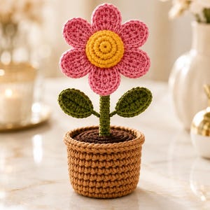Op de afbeelding: Een gehaakte bloem in een pot met een roze bloem met zes bloemblaadjes en een geel hart, een groene steel met twee blaadjes en een bruine bovenkant in een beige, getextureerde pot. Het object is volledig gemaakt van garen met zichtbare steken.