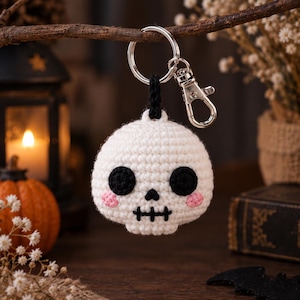 Puede incluir: Un llavero de calavera tejido a ganchillo en color blanco con grandes ojos circulares negros, una pequeña nariz triangular negra y una boca negra bordada. La calavera tiene pequeñas mejillas rosas tejidas y un anillo de metal plateado.