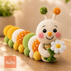 Op de afbeelding: Een gehaakt rupsspeeltje met een crèmekleurige kop, roze wangen en twee voelsprieten met groene en oranje bollen. Het lichaam bestaat uit afwisselende gele, witte, groene en oranje segmenten. Het houdt een witte margriet vast.