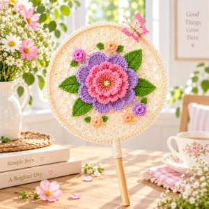 Puede incluir: Un abanico de mano redondo de ganchillo color crema con una flor grande y superpuesta de color rosa y morado en el centro, rodeada de flores de ganchillo más pequeñas en melocotón y morado, hojas verdes y un pequeño detalle de mariposa rosa.