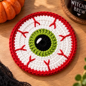 Puede incluir: Un posavasos redondo de ganchillo diseñado para parecerse a un globo ocular humano. El centro presenta una pupila negra con un reflejo blanco, rodeada por un iris verde brillante, una esclerótica blanca y un borde exterior rojo con venas.
