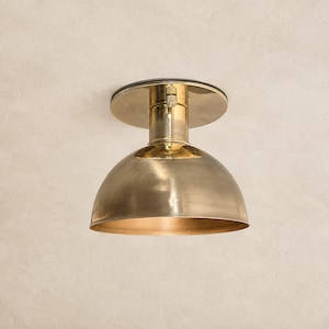 May include: A flush mount ceiling light fixture featuring a polished brass finish. The design consists of a circular mounting plate attached to a cylindrical neck that connects to a wide, dome-shaped shade with a smooth, metallic surface.