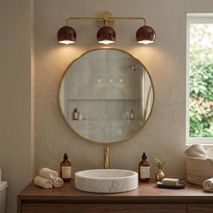 Könnte beinhalten: Ein Badezimmer-Waschtisch-Set mit einem runden Spiegel mit dünnem Goldrahmen, einem weißen Marmor-Aufsatzwaschbecken und einer dreiflammigen Wandleuchte mit dunkelroten Kugelschirmen und Goldfinish, montiert über einer hölzernen Arbeitsplatte.