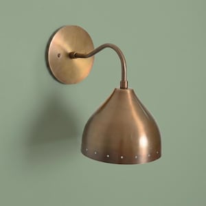 May include: A wall-mounted lamp featuring a circular brass backplate and a curved arm extending to a bell-shaped brass shade. The shade has a smooth metallic finish with a series of small, evenly spaced perforations along the bottom edge.