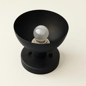 Può includere: Un'applique da parete nero opaco con un ampio paralume a forma di ciotola e una base circolare. L'apparecchio include un attacco centrale con una singola lampadina bianca smerigliata e due piccole manopole cilindriche sulla base.