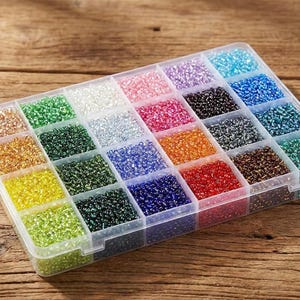 May include: A clear plastic rectangular storage container with twenty-four individual compartments filled with small, multi-coloured glass seed beads in shades of red, blue, green, yellow, orange, purple, pink, white, black, brown, and metallic tones.