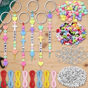 May include: A DIY keychain making kit featuring six assembled keychains with pastel heart and cross beads, personalized with letter cubes spelling EMAN, ARON, LON, TONY, MIYA, and GRACE, plus loose heart beads, cross beads, jump rings, and cord.
