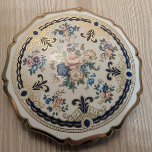 May include: A round compact mirror with a scalloped gold-tone metal edge. The top surface is white with a central floral bouquet in pink, blue, and green, surrounded by a decorative border of dark blue and gold filigree patterns and small dots.