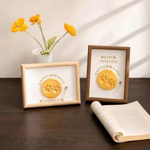 May include: Two wooden frames containing white inserts with gold text reading HAPPY 100 DAYS and BABY 20260305. Each frame features a circular yellow clay disc with two paw prints and a small glass vial. Yellow flowers in a white vase are nearby.
