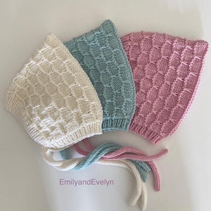 May include: Three knitted baby bonnets in white, light blue and pink with matching ties. The bonnets have a textured stitch pattern.