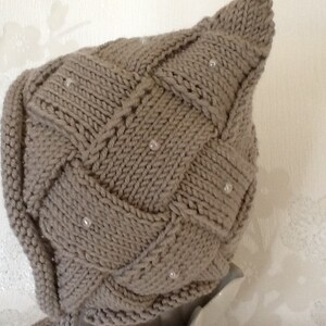 May include: A knitted, taupe-colored hat with a woven design and small, pearl-like embellishments. The hat has a pointed top and a snug fit, suitable for cold weather. The texture of the knitwear is visible, highlighting the craftsmanship.