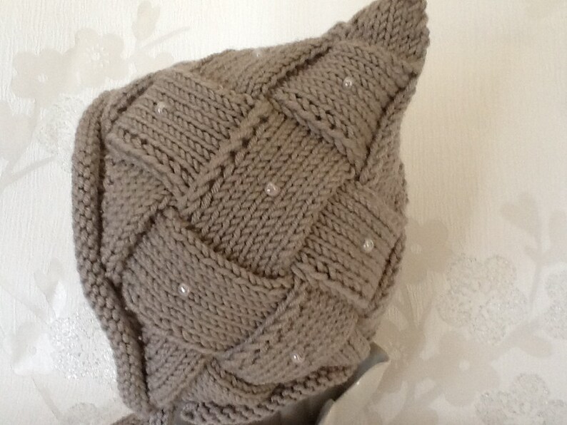 May include: A knitted, taupe-colored hat with a woven design and small, pearl-like embellishments. The hat has a pointed top and a snug fit, suitable for cold weather. The texture of the knitwear is visible, highlighting the craftsmanship.
