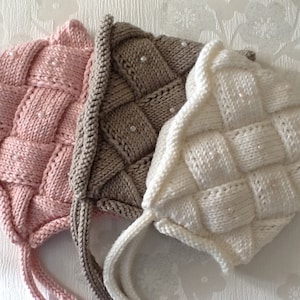 May include: Three knitted baby bonnets in pink, tan, and white, each with a woven pattern and adorned with small, pearlescent beads. The bonnets have ties for securing under the chin.
