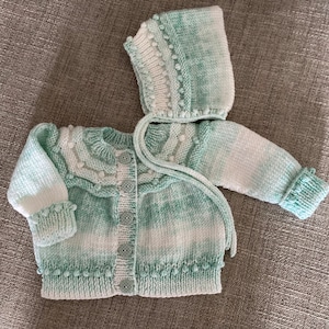 May include: A green and white knitted baby cardigan with a matching hooded hat. The cardigan has a button closure and a cable knit pattern. The hat has a pom-pom on top.