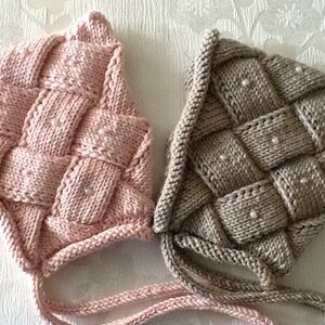 May include: Two knitted baby bonnets. One is light pink, the other is a neutral beige color. Both bonnets have a woven design with small white beads. The bonnets have ties to secure them under the chin.
