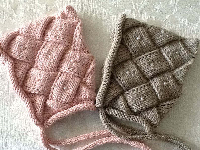 May include: Two knitted baby bonnets. One is light pink, the other is a neutral beige color. Both bonnets have a woven design with small white beads. The bonnets have ties to secure them under the chin.