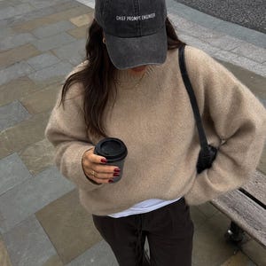 May include: A charcoal gray baseball cap with the text CHIEF PROMPT ENGINEER in white, a beige long-sleeve knit sweater, a white undershirt, and dark brown trousers. A black shoulder bag and a black coffee cup are also visible in the image.
