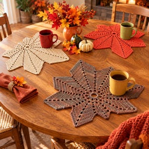 May include: Three crocheted leaf-shaped placemats in beige, orange, and brown, each featuring a central circular opening and radiating stitch patterns. The set includes a red mug, a green mug, and a yellow mug, alongside a folded orange napkin.